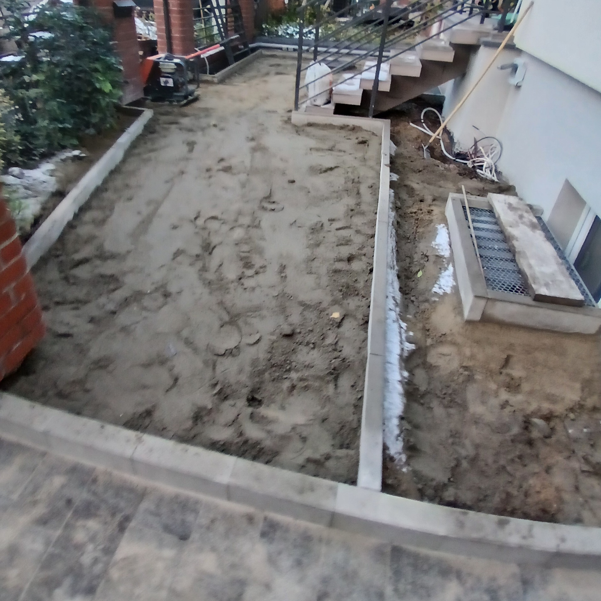 Przygotowany teren pod budowę tarasu: wyrównana ziemia otoczona betonowymi obrzeżami, z widoczną ubijarką w tle i schodami prowadzącymi do piwnicy.