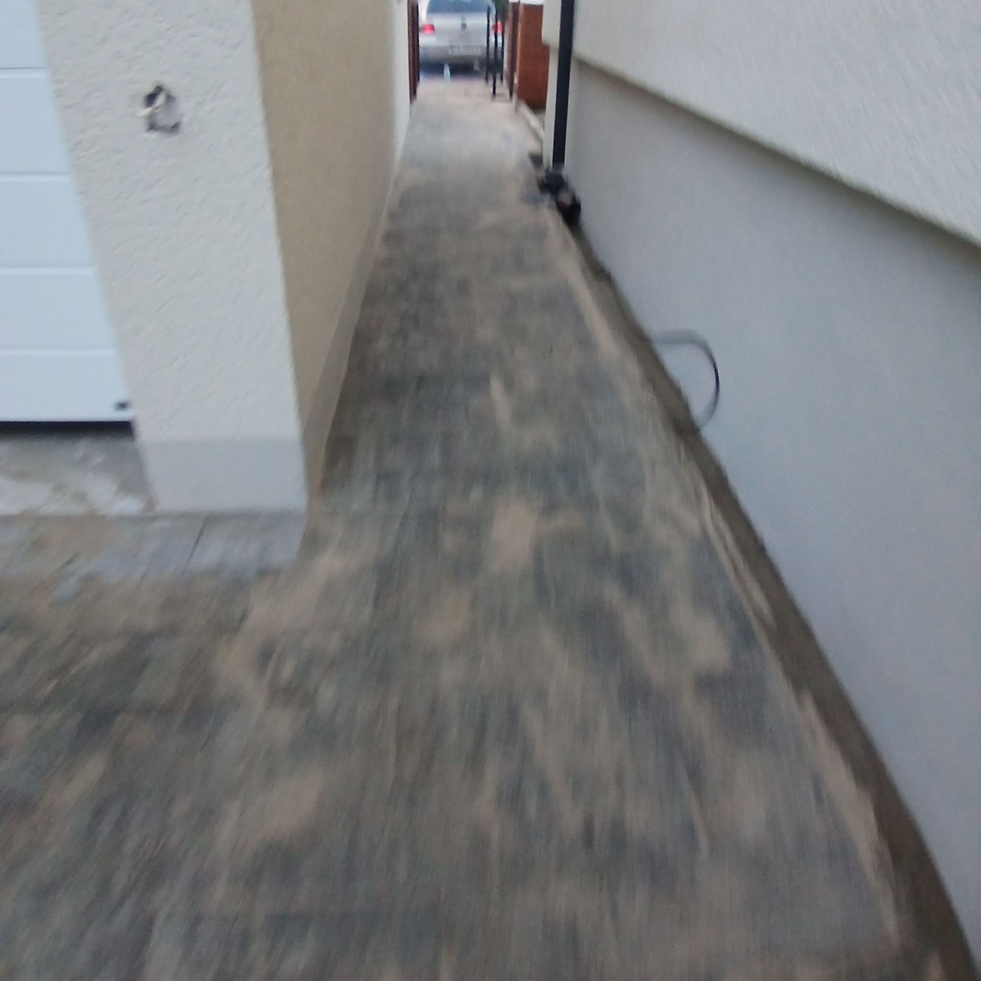 Ułożona szara kostka brukowa na wąskim chodniku między dwoma budynkami, posypana piaskiem, w tle zaparkowany samochód.