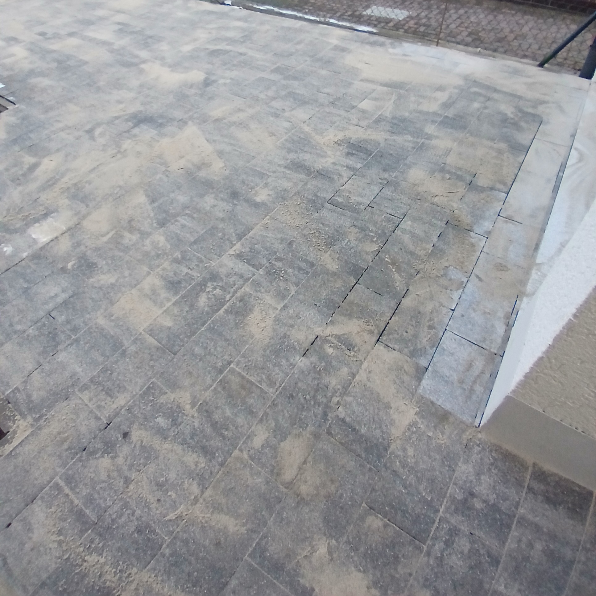 Ułożona szara kostka brukowa w trakcie czyszczenia, widoczne ślady piasku na powierzchni, krawędź schodów w tle.