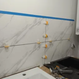 Montaż płytek imitujących marmur na ścianie nad zlewem w kuchni, widoczne klipsy poziomujące, niebieska taśma malarska i piec gazowy Termet.