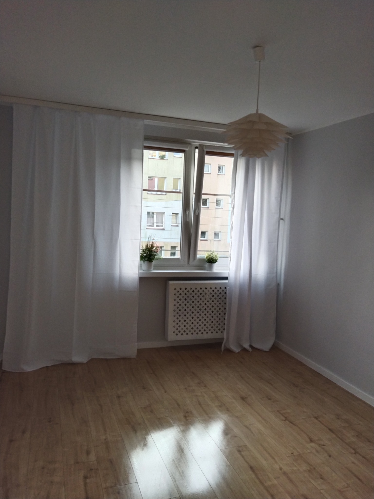 Jasne wnętrze pokoju z drewnianą podłogą, białymi zasłonami przy oknie z widokiem na blok, grzejnikiem pod parapetem i lampą w kształcie kwiatu zwisającą z sufitu.
