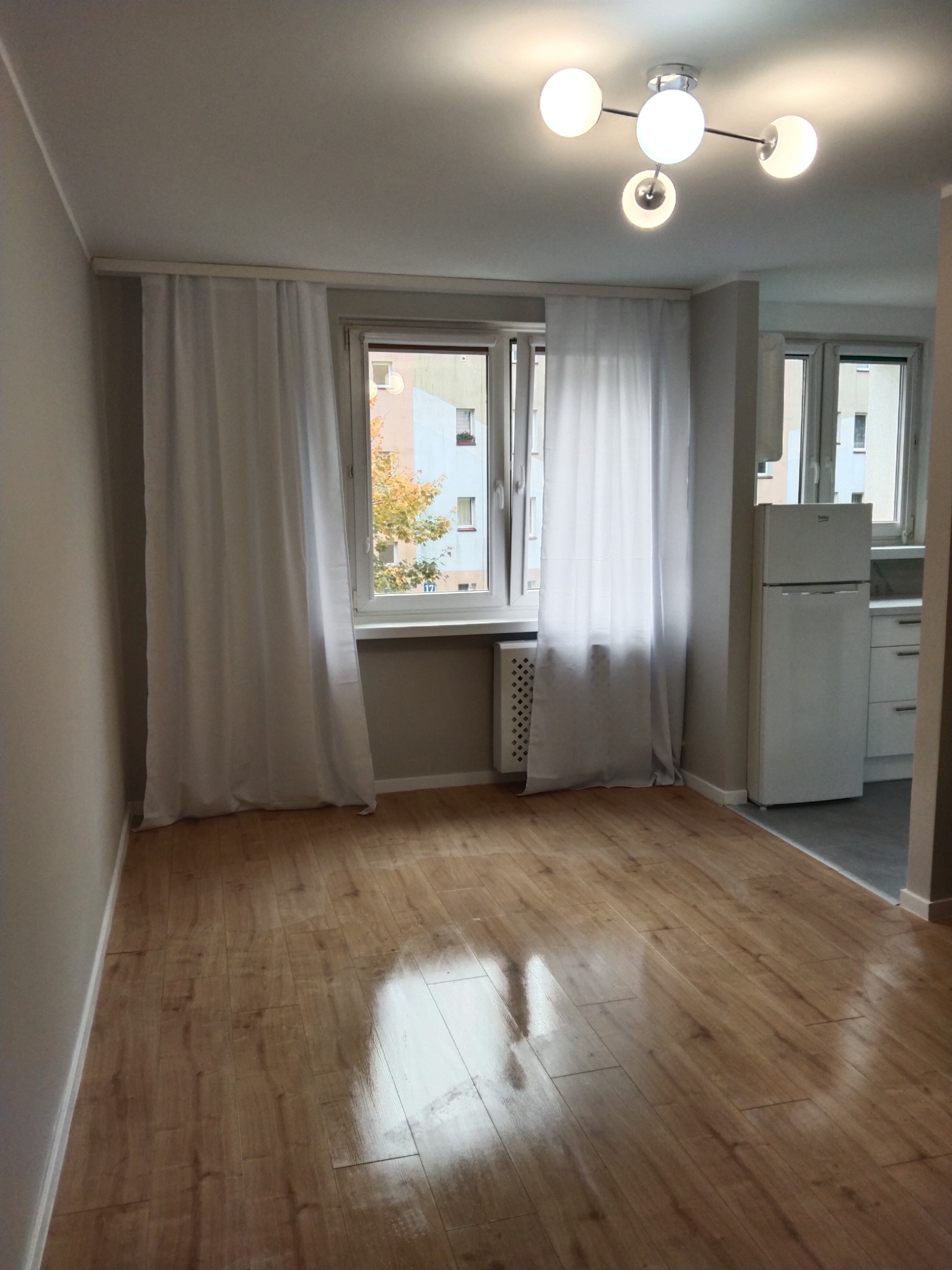 Jasne wnętrze pokoju z panelami podłogowymi, białymi zasłonami przy oknach i nowoczesną lampą sufitową z kulistymi kloszami. Widoczny fragment kuchni z białą lodówką.