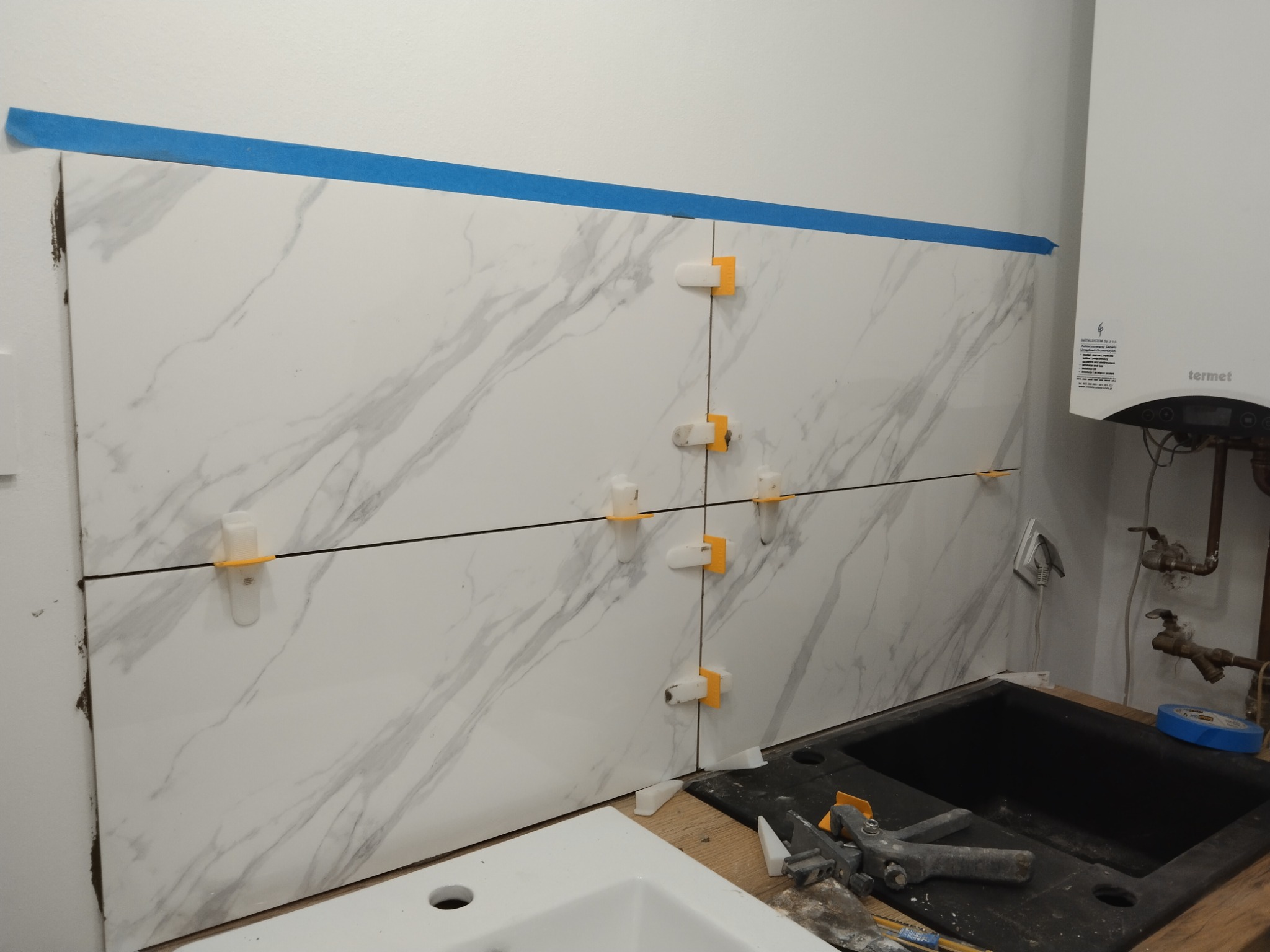 Montaż płytek imitujących marmur na ścianie nad zlewem w kuchni, widoczne klipsy poziomujące, niebieska taśma malarska i piec gazowy Termet.