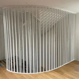 Biała, zaokrąglona balustrada z pionowych prętów zabezpieczająca schody, częściowo owinięta folią ochronną podczas montażu, widoczny fragment drewnianej podłogi i przygotowania do wykończenia ściany.