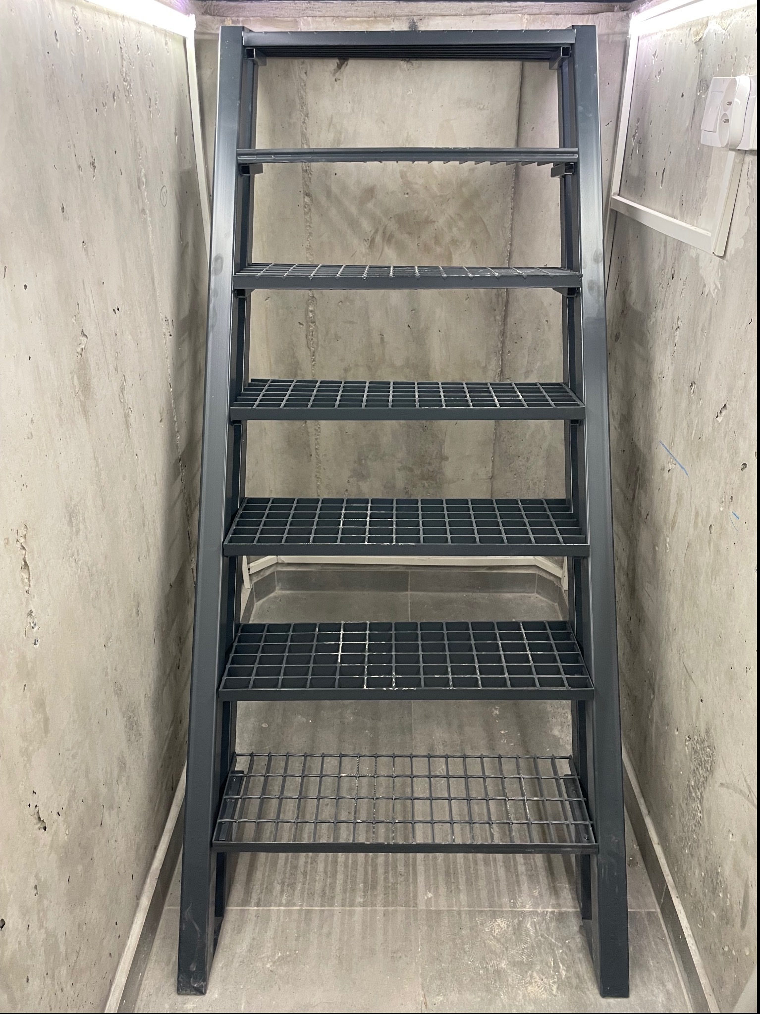 Metalowa drabina schodkowa z kratowanymi stopniami, umieszczona w surowym, betonowym wnętrzu z widocznym oświetleniem LED u góry.