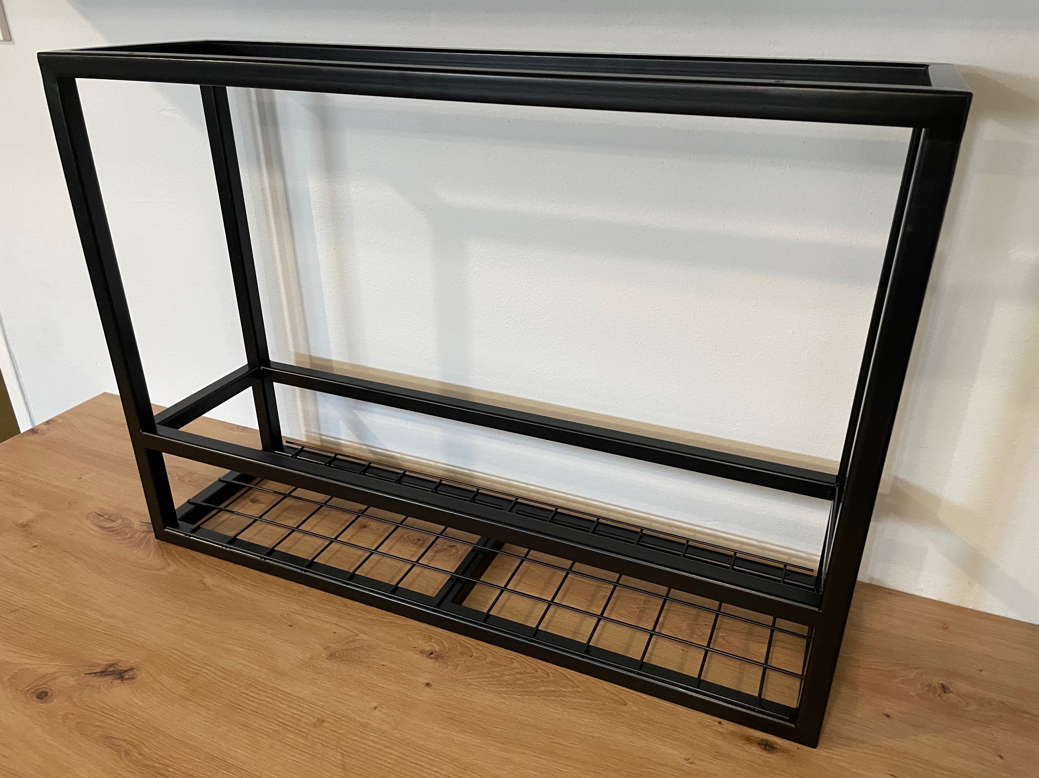 Czarny, metalowy stelaż z kratką na dole, prawdopodobnie na buty, ustawiony na drewnianej podłodze przy białej ścianie.