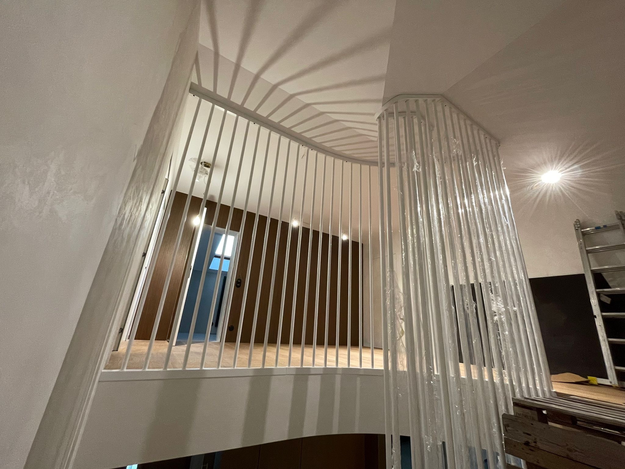 Wewnętrzna balustrada schodowa z białych metalowych prętów, częściowo owinięta folią ochronną, z widocznym fragmentem drewnianej podłogi i ścian w tle.