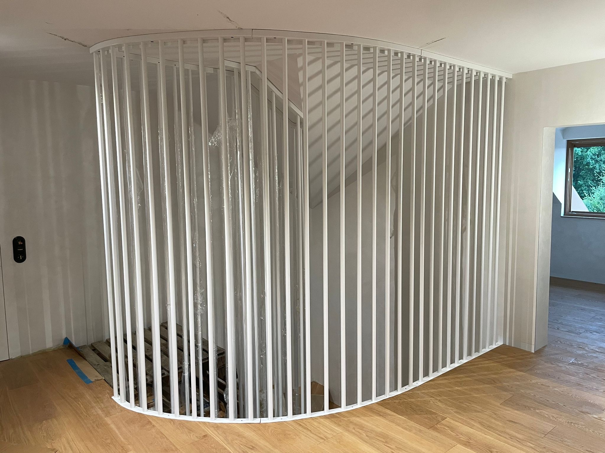Biała, zaokrąglona balustrada z pionowych prętów zabezpieczająca schody, częściowo owinięta folią ochronną podczas montażu, widoczny fragment drewnianej podłogi i przygotowania do wykończenia ściany.