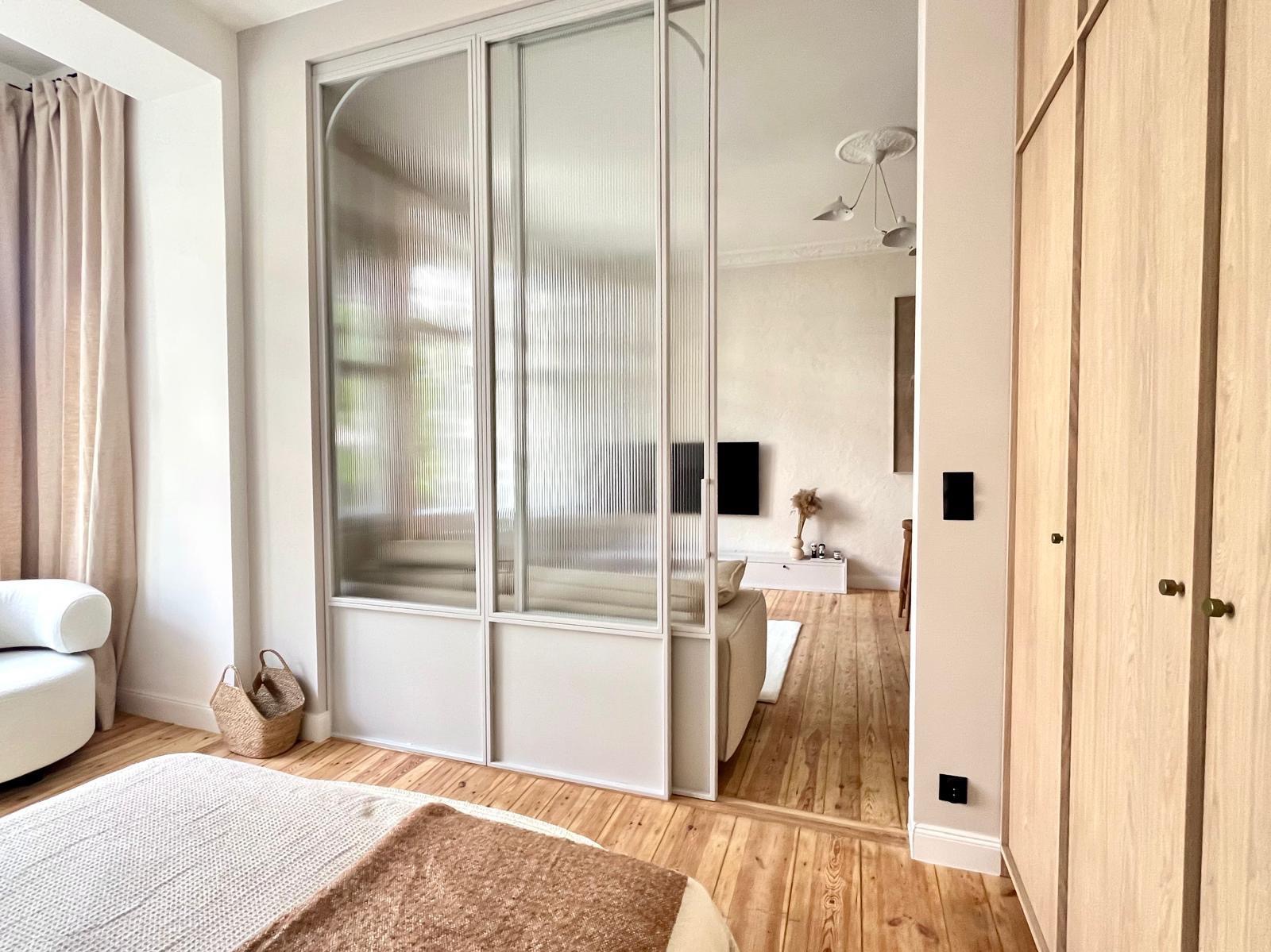 Przesuwne drzwi w metalowej ramie z ryflowanym szkłem oddzielające sypialnię od salonu, widok z perspektywy łóżka, drewniana podłoga, jasne wnętrze.