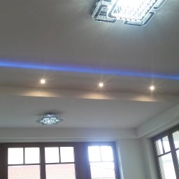 Krystian Gula - Wykończony sufit w pokoju z oświetleniem LED w kolorze niebieskim, halogenami i dwoma kryształowymi lampami, widok na okna z ciemnymi ramami.