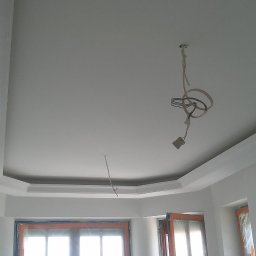 Krystian Gula - Pomieszczenie po malowaniu na biało, widoczne listwy dekoracyjne pod sufitem i wystające kable elektryczne.