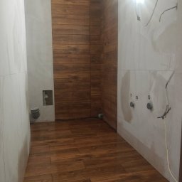 Krystian Gula - Wnętrze łazienki w trakcie remontu z drewnopodobnymi płytkami na podłodze i ścianach, widoczne przygotowane punkty instalacyjne na ścianie, surowe wykończenie.
