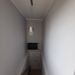 Krystian Gula - Klatka schodowa w szarościach z wiszącą lampą kryształową i czarnym grzejnikiem. Ściany pomalowane na jednolity, jasny odcień szarości.