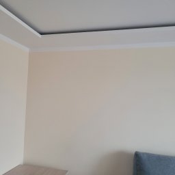 Krystian Gula - Jasny pokój po odświeżeniu: kremowe ściany z białym, ozdobnym pasem pod sufitem, widoczny fragment mebla i niebieskiego obicia.