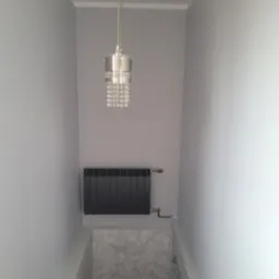 Wnętrze pomalowane na szaro z grzejnikiem panelowym i lampą wiszącą z kryształkami, widoczna dekoracyjna faktura ściany w dolnej części.