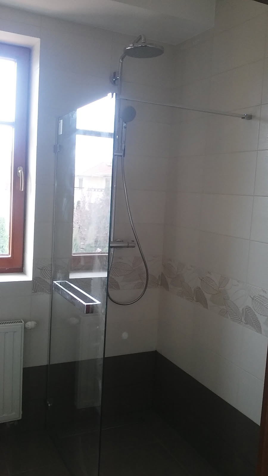 Nowoczesna kabina prysznicowa ze szklanymi drzwiami, deszczownicą i panelem z dekoracyjnymi płytkami w łazience z oknem.