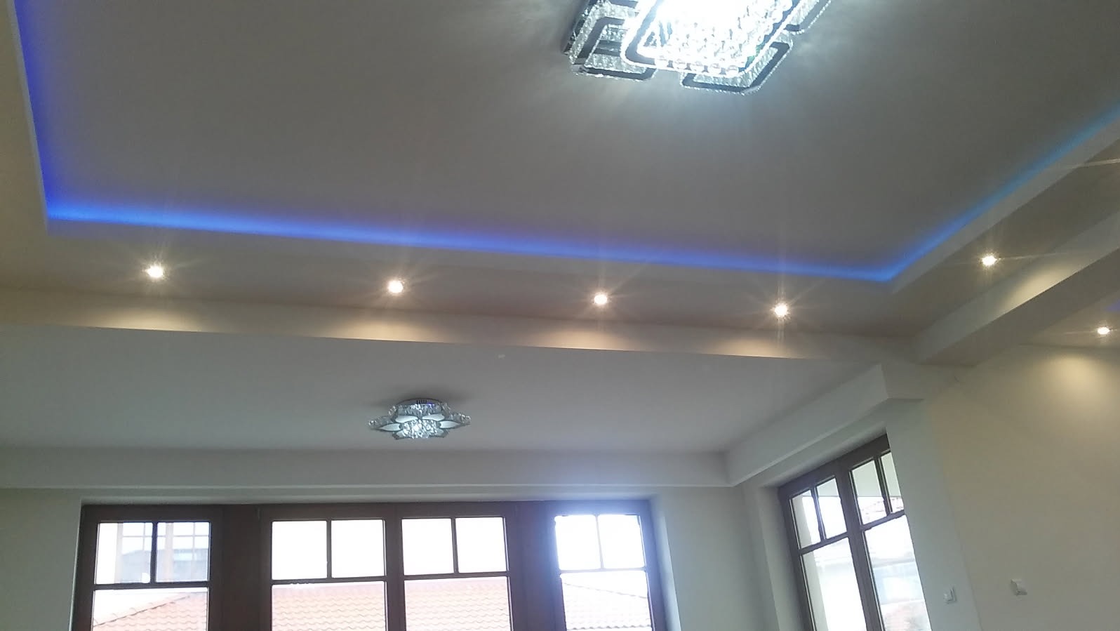 Wykończony sufit w pokoju z oświetleniem LED w kolorze niebieskim, halogenami i dwoma kryształowymi lampami, widok na okna z ciemnymi ramami.