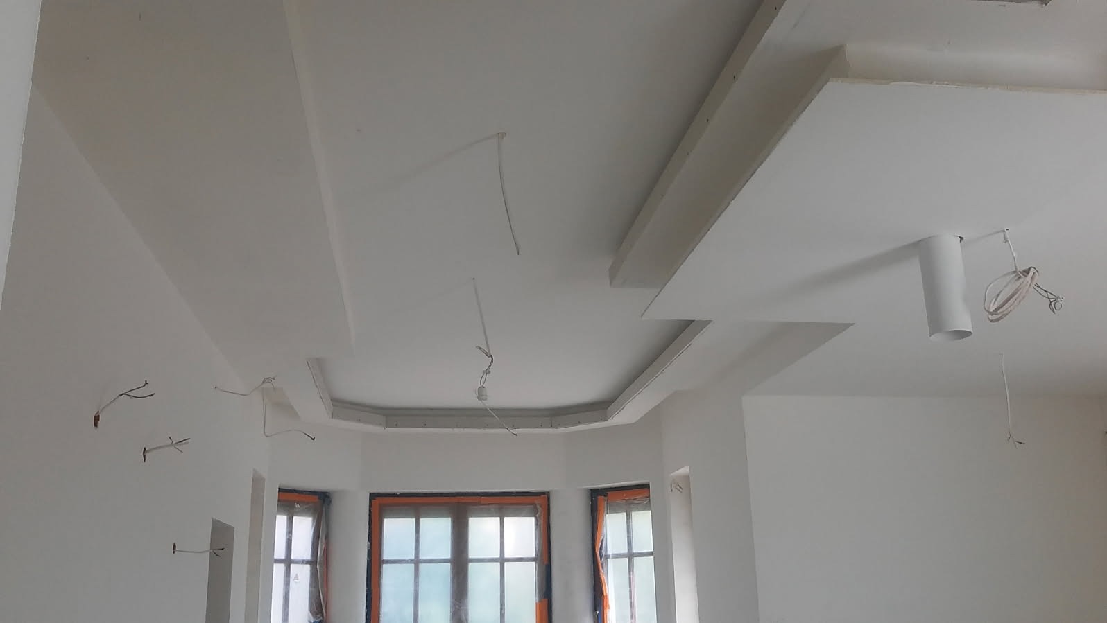 Wnętrze w trakcie prac wykończeniowych: widoczne fragmenty sufitu z elementami dekoracyjnymi z płyt gipsowo-kartonowych, wystające przewody elektryczne oraz okna zabezpieczone folią malarską.