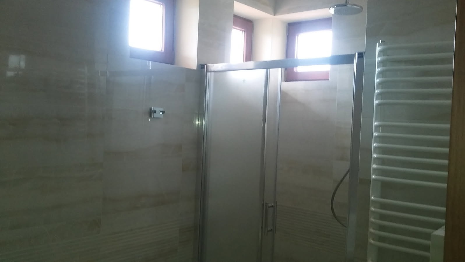 Wykończona łazienka z kabiną prysznicową ze szklanymi drzwiami, grzejnikiem drabinkowym i beżowymi płytkami imitującymi marmur na ścianach.