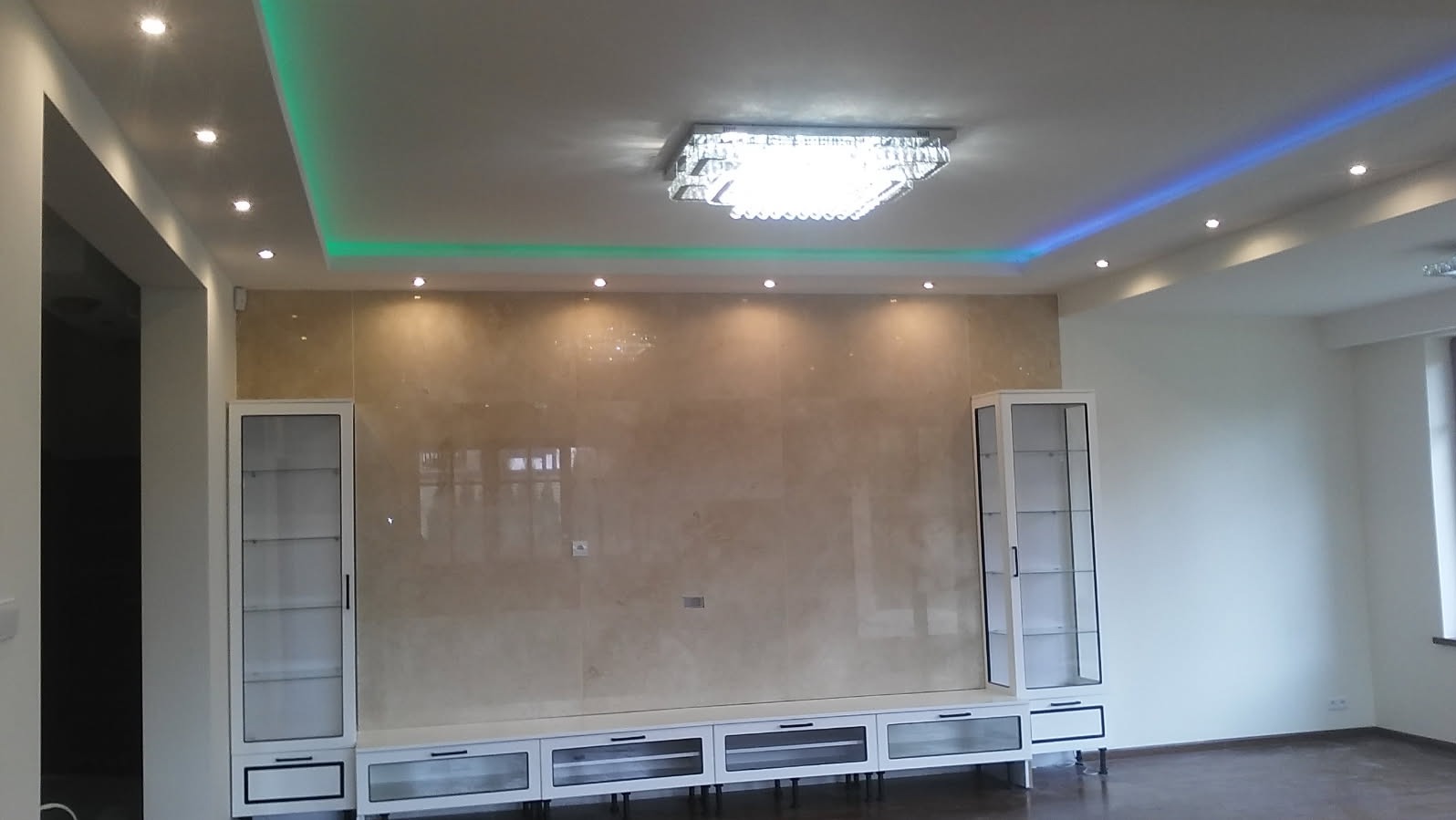 Eleganckie wnętrze salonu po remoncie: ściana wykończona płytami imitującymi marmur, podświetlany sufit z kryształowym żyrandolem, białe przeszklone regały po bokach.