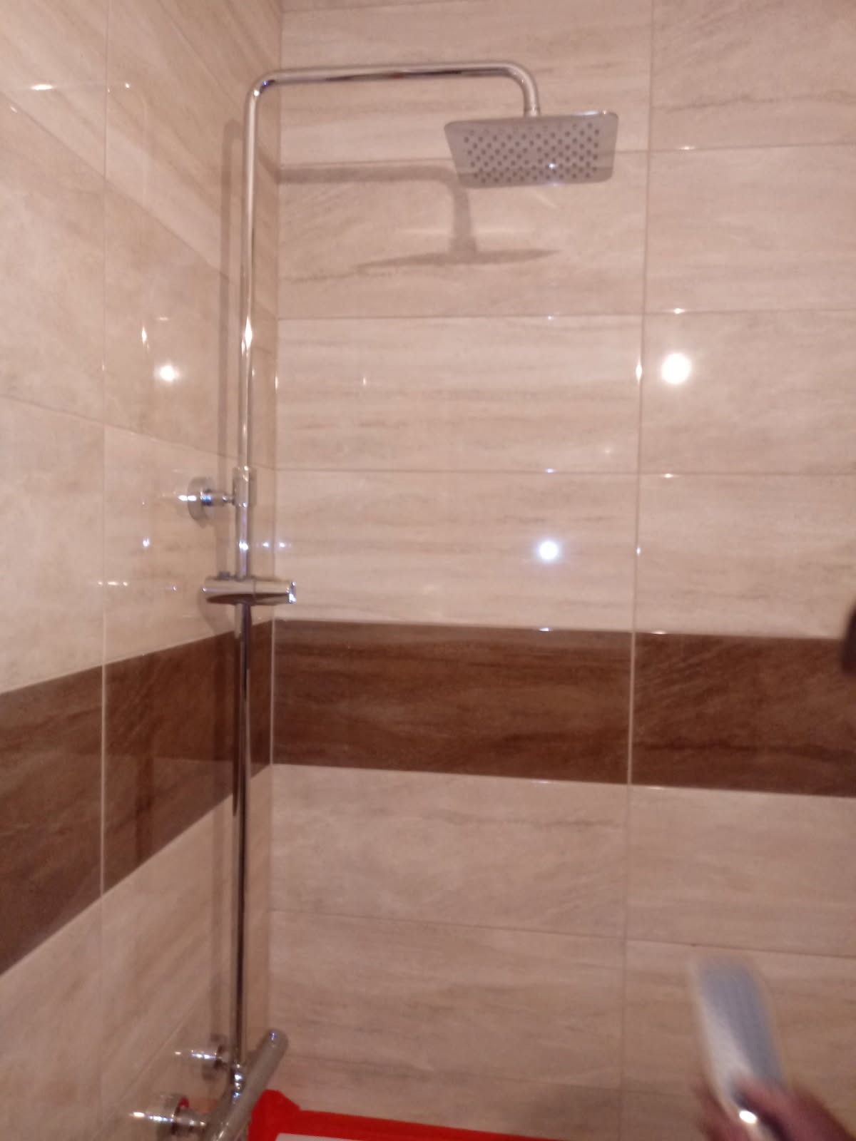 Zamontowana chromowana kolumna natryskowa z deszczownicą na tle beżowych płytek z brązowym pasem w nowej łazience.