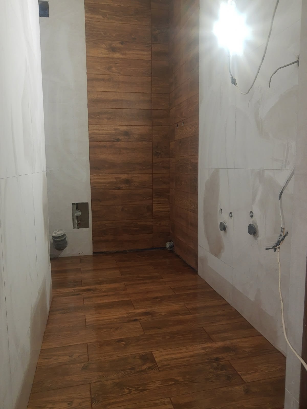 Wnętrze łazienki w trakcie remontu z drewnopodobnymi płytkami na podłodze i ścianach, widoczne przygotowane punkty instalacyjne na ścianie, surowe wykończenie.