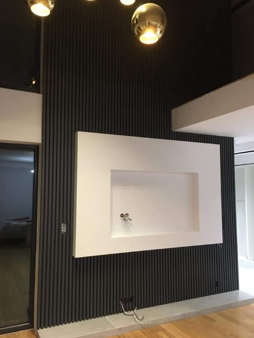 Nowoczesna ściana z ciemnymi, pionowymi panelami i białą wnęką na telewizor z wystającymi kablami, oświetlona złotymi lampami sufitowymi.