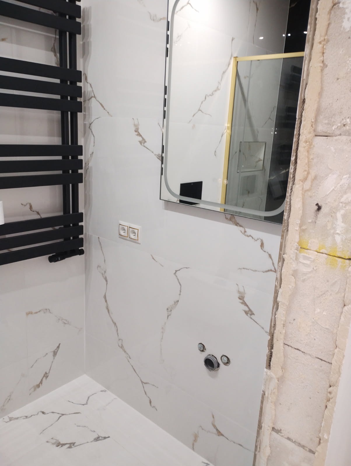 Fragment łazienki w trakcie wykańczania: białe płytki z marmurkowym wzorem, czarny grzejnik drabinkowy, lustro z podświetleniem LED i fragment surowej ściany z widoczną instalacją sanitarną.