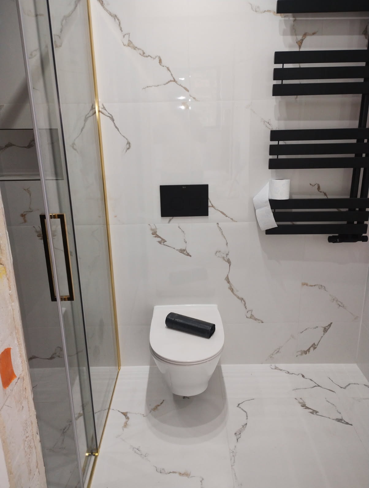 Nowoczesna łazienka z białymi płytkami imitującymi marmur, czarnym grzejnikiem drabinkowym, podwieszaną toaletą z czarnym przyciskiem i kabiną prysznicową ze złotymi profilami. Na desce toalety...