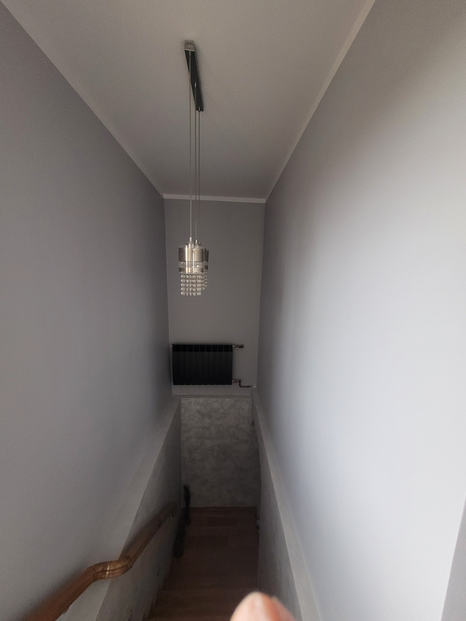 Klatka schodowa w szarościach z wiszącą lampą kryształową i czarnym grzejnikiem. Ściany pomalowane na jednolity, jasny odcień szarości.