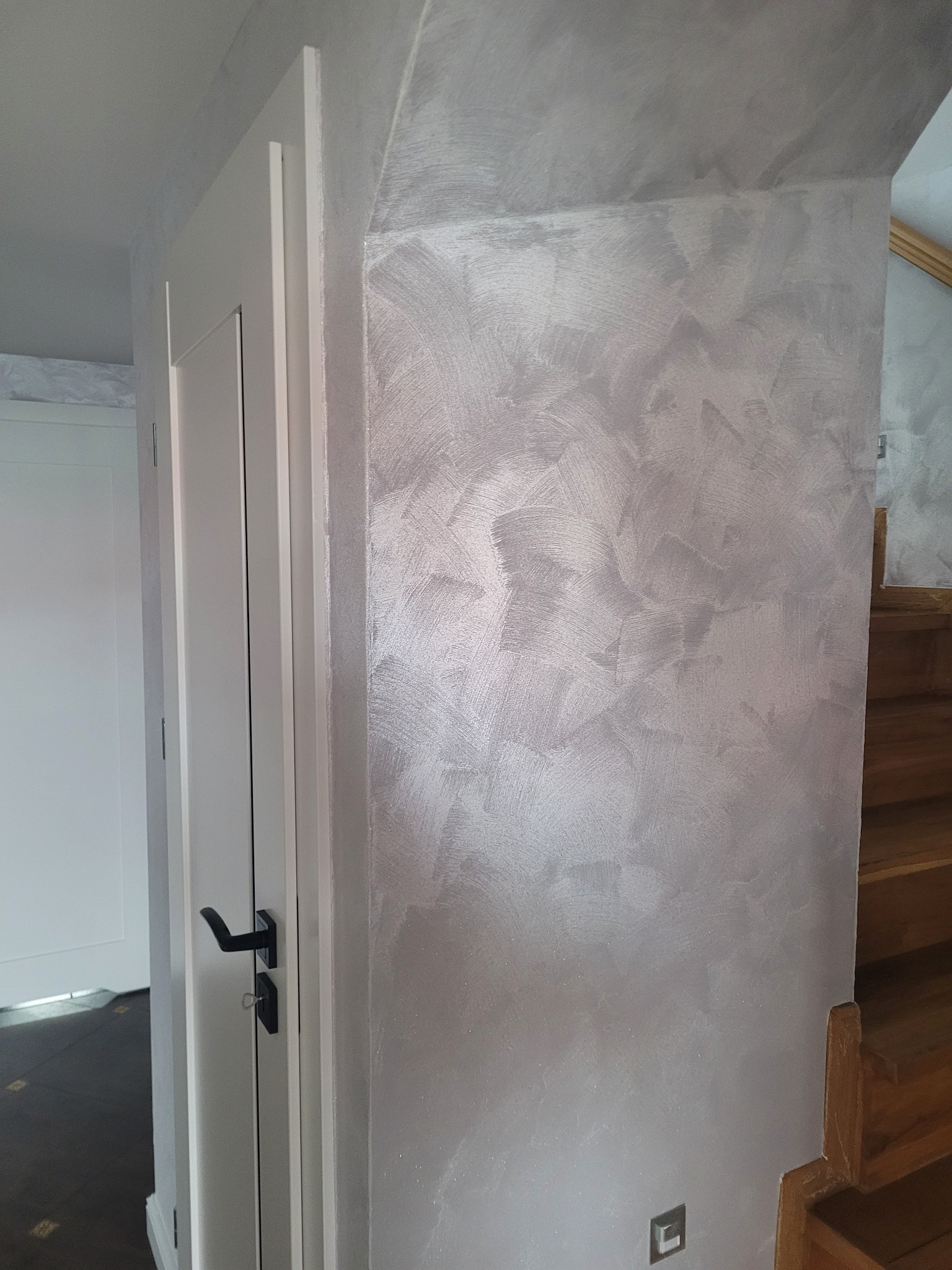 Ściana wewnętrzna z dekoracyjną, srebrzystą fakturą, widoczna obok białych drzwi z czarną klamką i fragmentu drewnianych schodów.