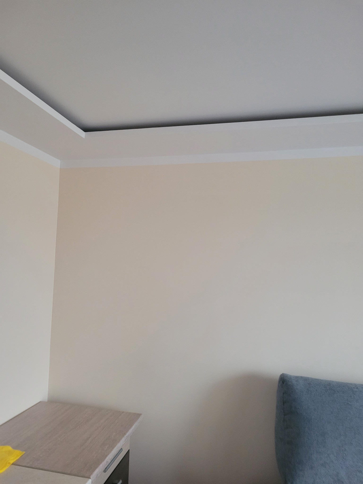 Jasny pokój po odświeżeniu: kremowe ściany z białym, ozdobnym pasem pod sufitem, widoczny fragment mebla i niebieskiego obicia.