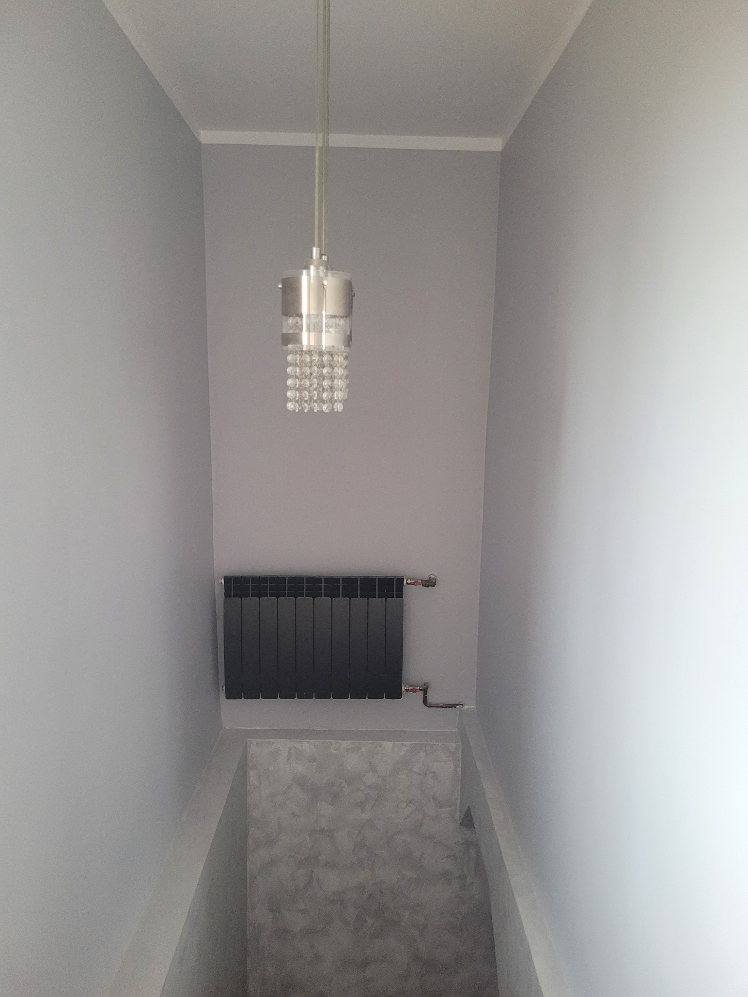 Wnętrze pomalowane na szaro z grzejnikiem panelowym i lampą wiszącą z kryształkami, widoczna dekoracyjna faktura ściany w dolnej części.