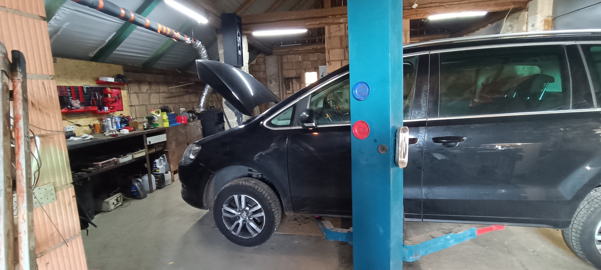 Czarny minivan na podnośniku w warsztacie, z otwartą maską, widoczne narzędzia i wyposażenie warsztatowe.