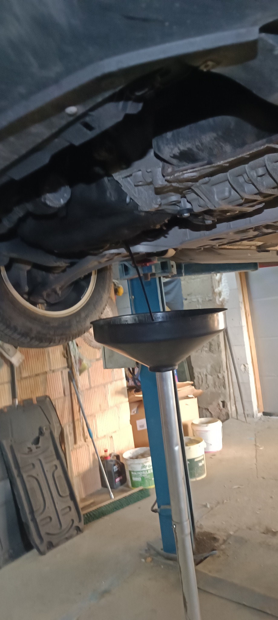 Ciemny olej silnikowy spływa z podwozia samochodu uniesionego na podnośniku do czarnego lejka na stojaku. Widoczny fragment koła, elementy zawieszenia, podwozie, ceglana ściana w tle oraz narzędzia...