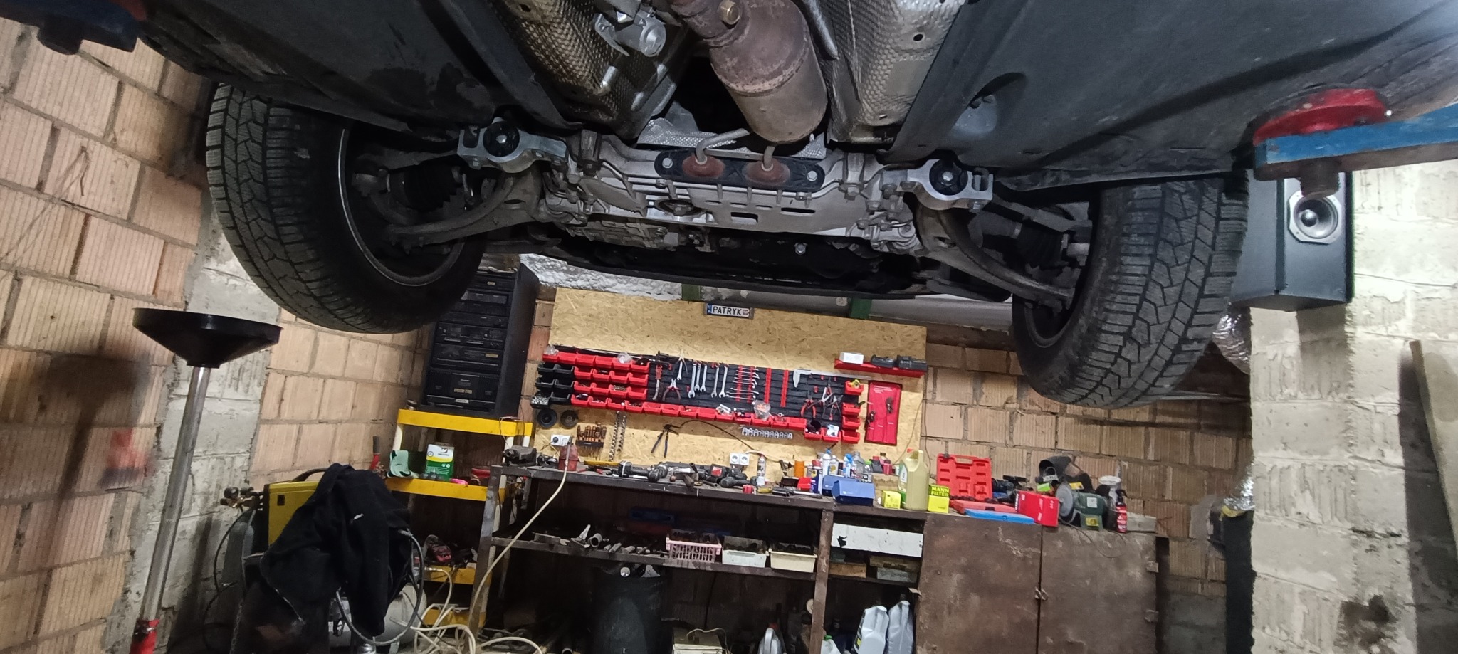 Podwozie samochodu na podnośniku w warsztacie, widoczny układ wydechowy, elementy zawieszenia, koła z oponami zimowymi, narzędzia na ścianie i pojemnik na olej pod spodem.