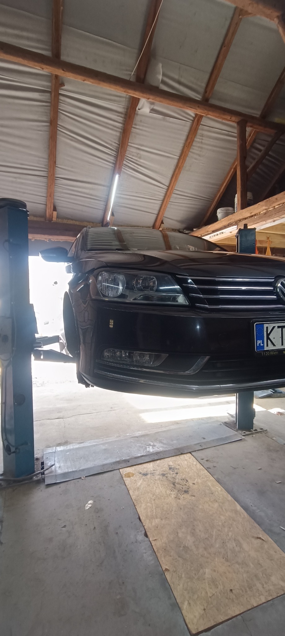 Czarny samochód osobowy na podnośniku w warsztacie, widok z dołu na przednią część pojazdu, widoczne oświetlenie i drewniana konstrukcja dachu.