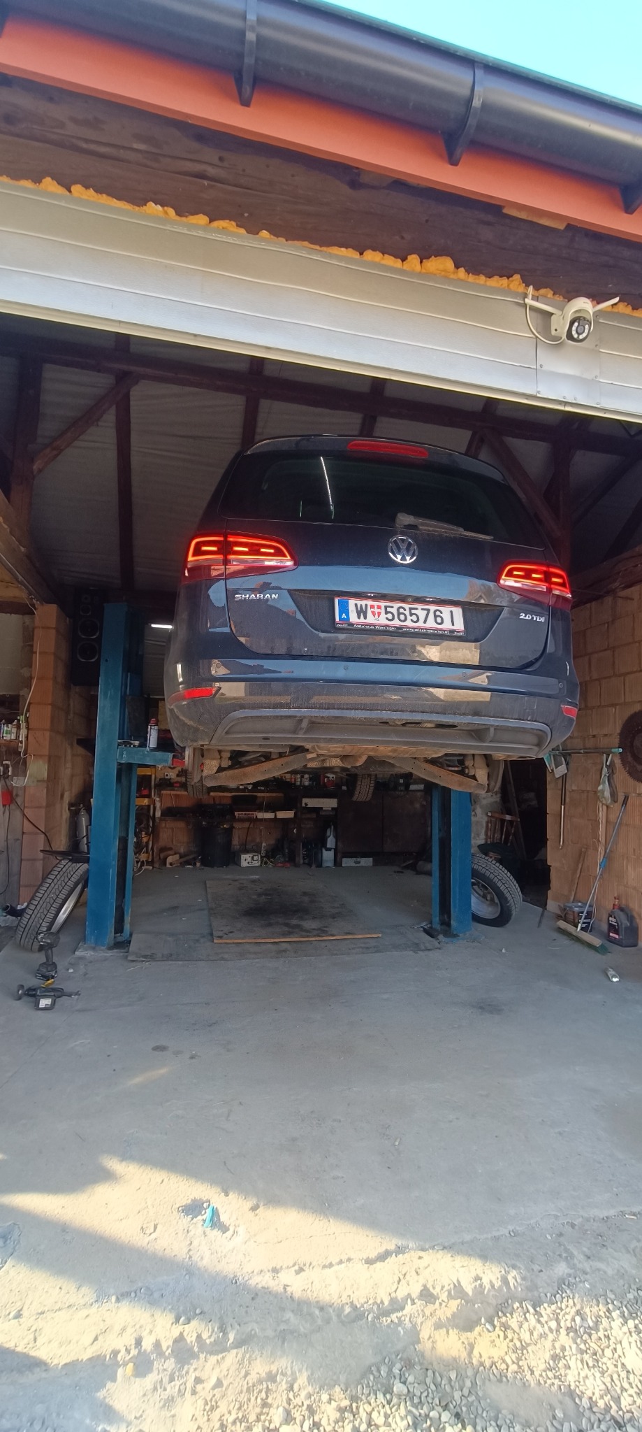 Samochód marki Volkswagen Sharan na podnośniku w warsztacie, widoczny numer rejestracyjny Austrii, oświetlenie warsztatowe i elementy konstrukcyjne budynku.