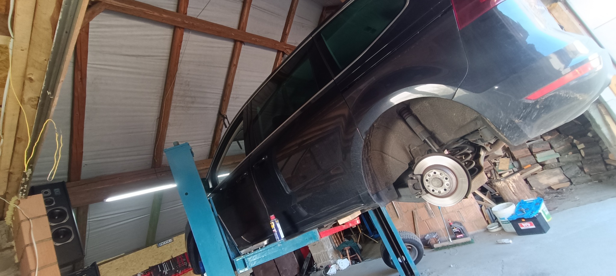 Czarny samochód na podnośniku warsztatowym z widocznym zdjętym kołem i tarczą hamulcową, w tle sterta drewna i narzędzia.