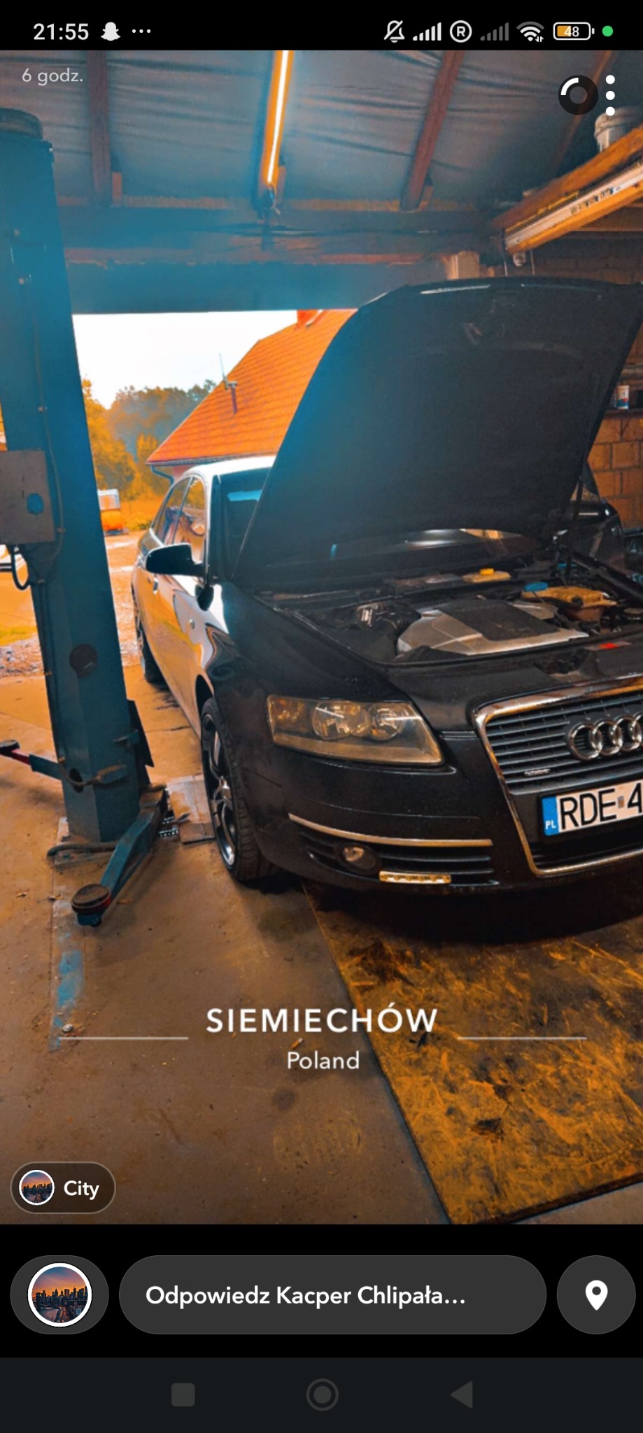 Czarny samochód Audi z otwartą maską, widoczny silnik, na podnośniku w warsztacie, podłoga z płyty OSB, oświetlenie warsztatowe.