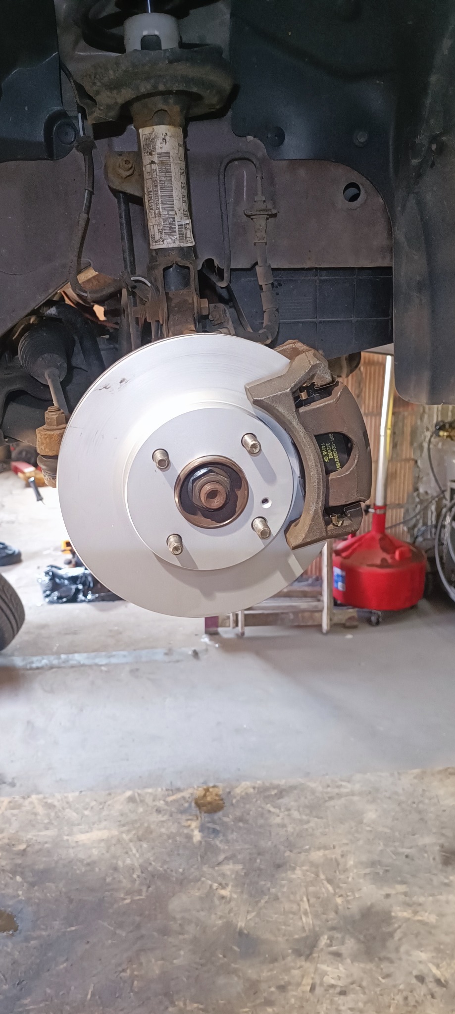 Nowa, srebrna tarcza hamulcowa z zaciskiem zamontowana w samochodzie, widoczne elementy zawieszenia i przewody hamulcowe, w tle warsztat z czerwonym kanistrem.