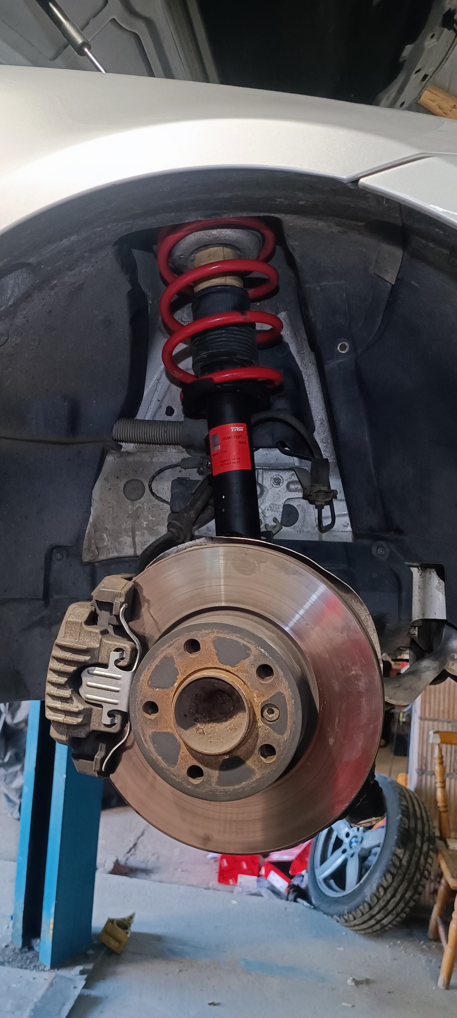 Zawieszenie samochodu na podnośniku, widoczny amortyzator z czerwoną sprężyną, tarcza hamulcowa i zacisk hamulcowy, koło w tle.