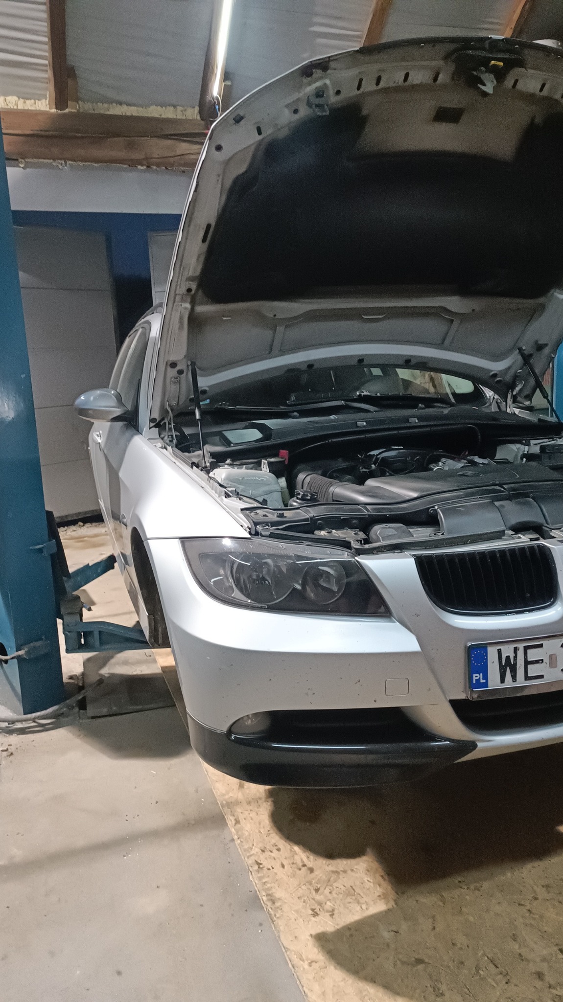 Srebrny samochód marki BMW na podnośniku w warsztacie, z otwartą maską i widocznym silnikiem, oświetlony lampą sufitową.