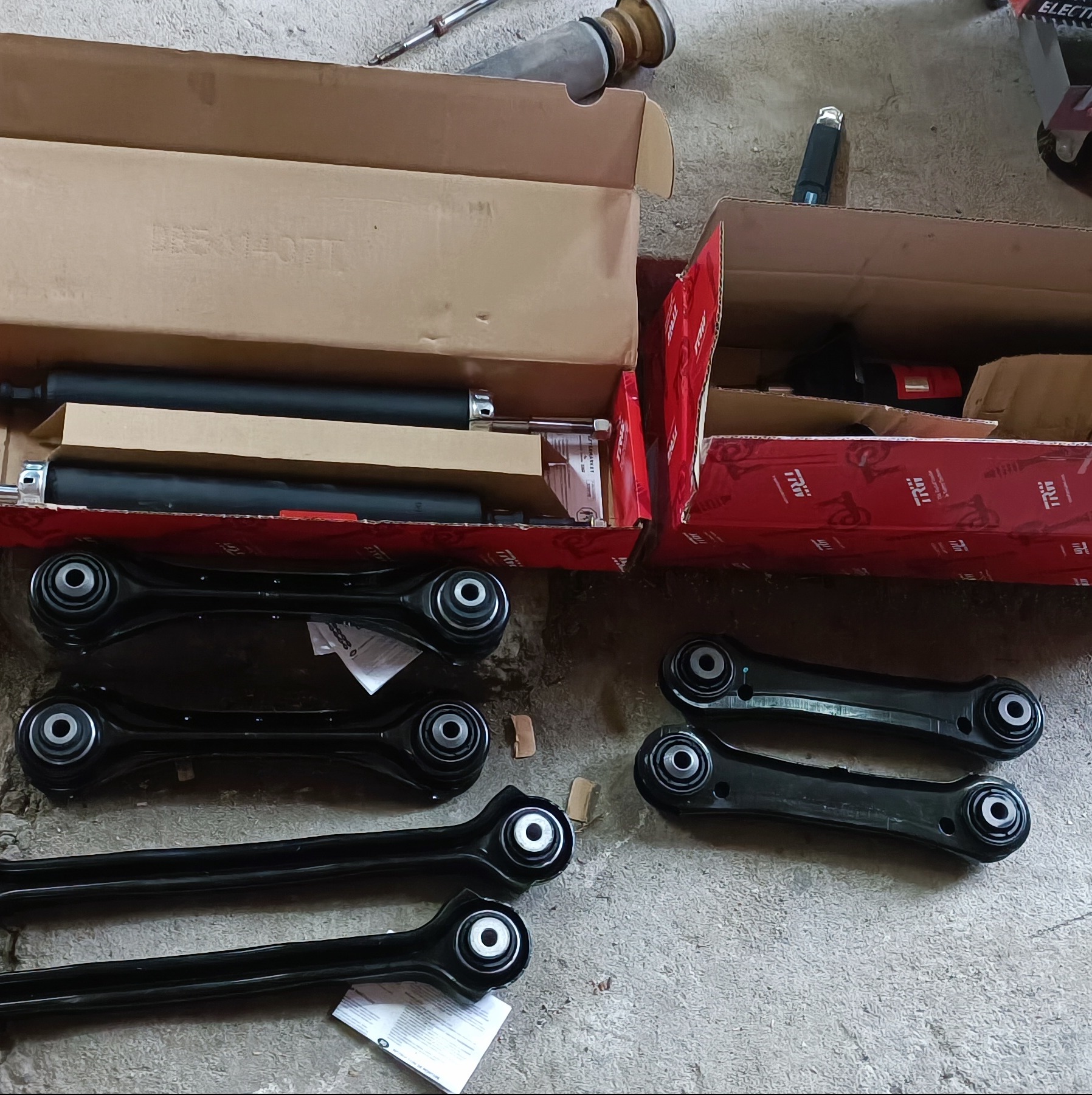 Nowe wahacze samochodowe i amortyzatory w pudełkach, gotowe do montażu, leżące na betonowej podłodze warsztatu, widoczne narzędzia i akcesoria.