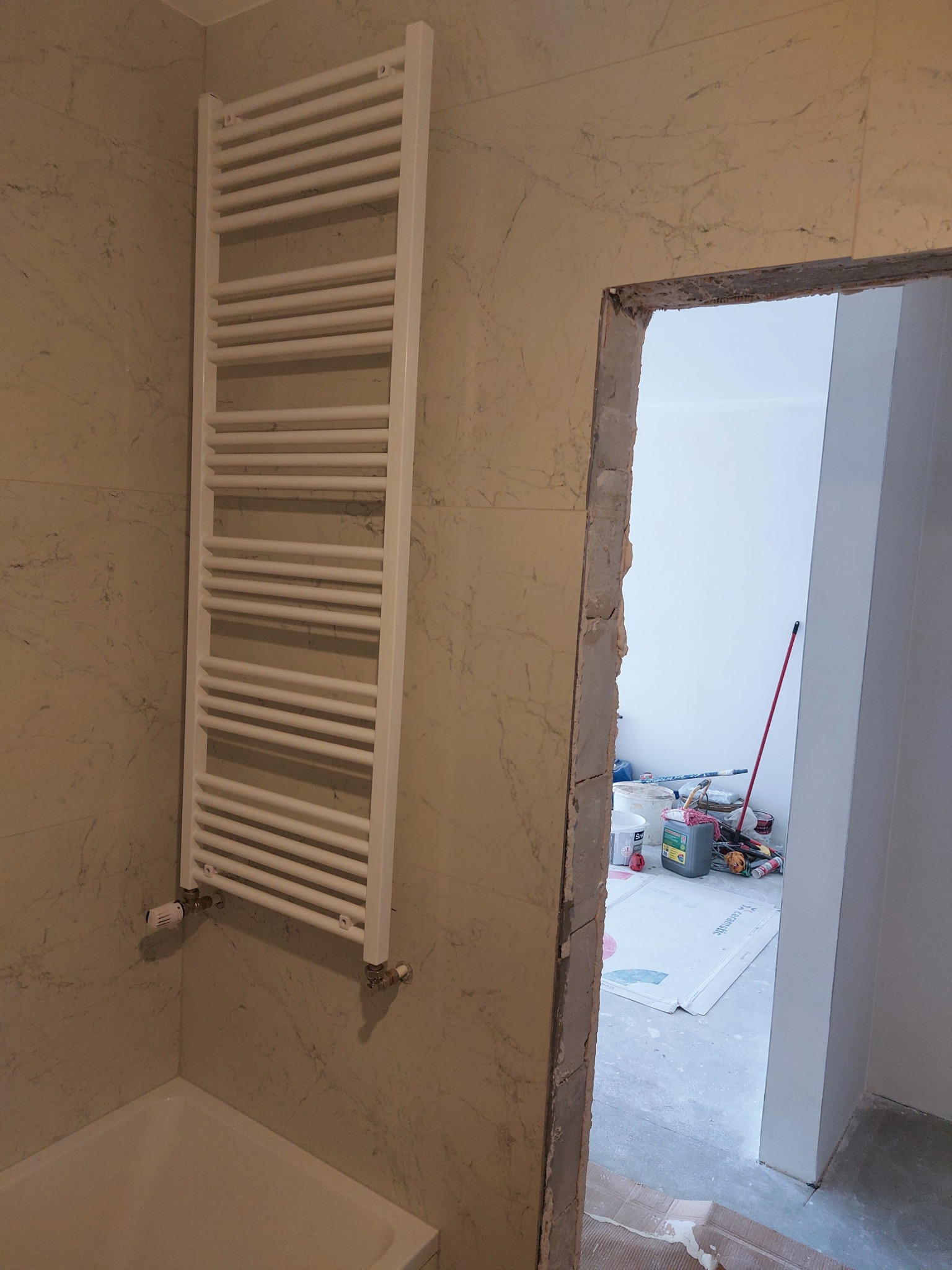 Biały grzejnik drabinkowy zamontowany na ścianie z marmurową fakturą w łazience. Widoczne materiały budowlane i narzędzia przez otwarte drzwi.