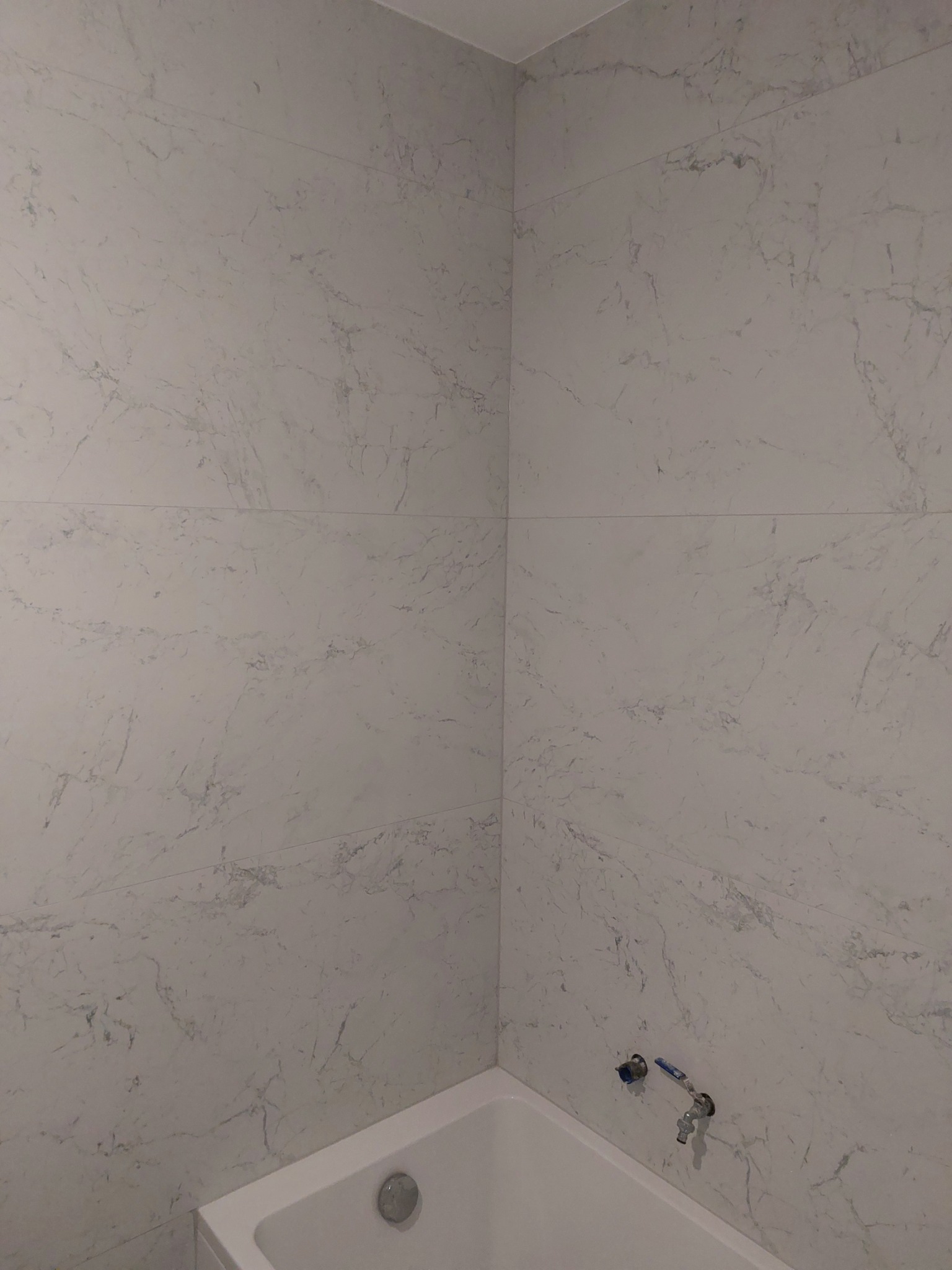 Wykończona ściana w łazience z białymi płytkami imitującymi marmur nad wanną. Widoczne elementy instalacji wodnej.