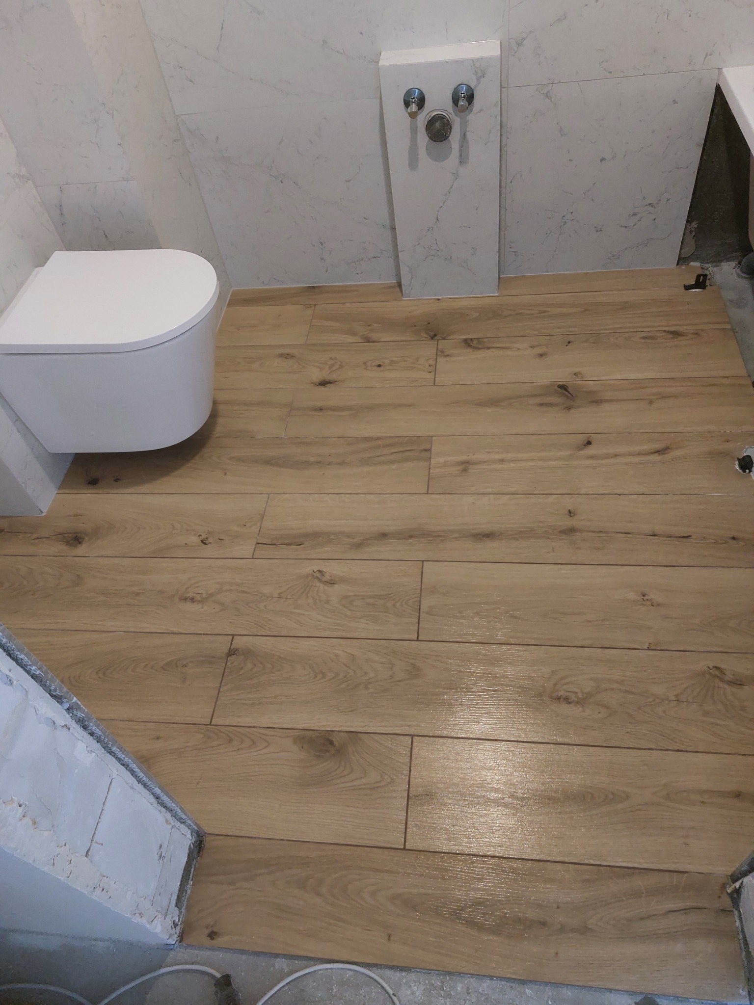 Ułożona podłoga z paneli imitujących drewno w łazience z białymi płytkami marmurkowymi na ścianach i podwieszaną toaletą. Widoczny fragment instalacji wodnej.