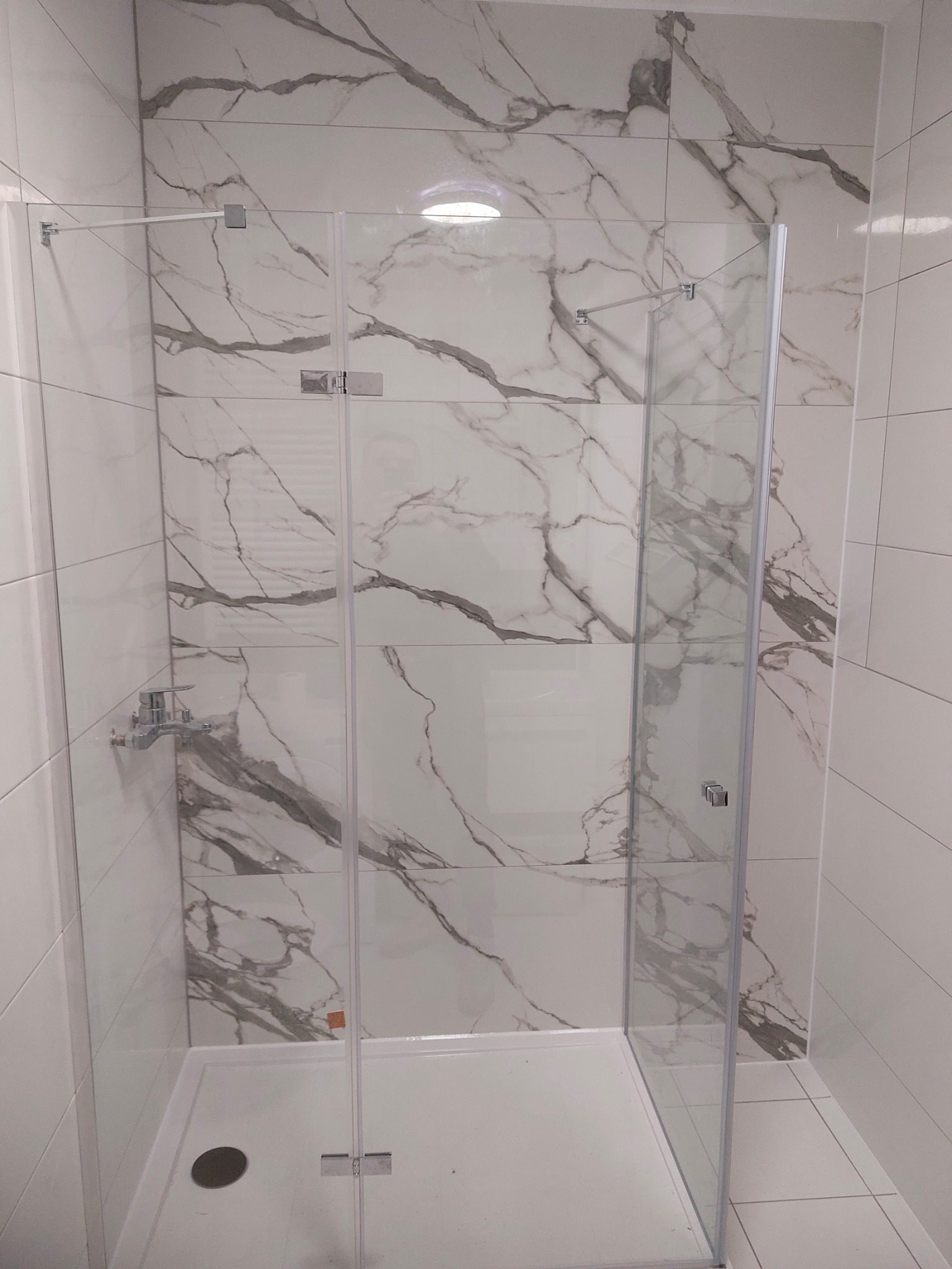 Kabina prysznicowa z marmurowymi płytkami, chromowana bateria i odpływ liniowy. Nowoczesna łazienka po remoncie, jasne wnętrze, minimalistyczny design.