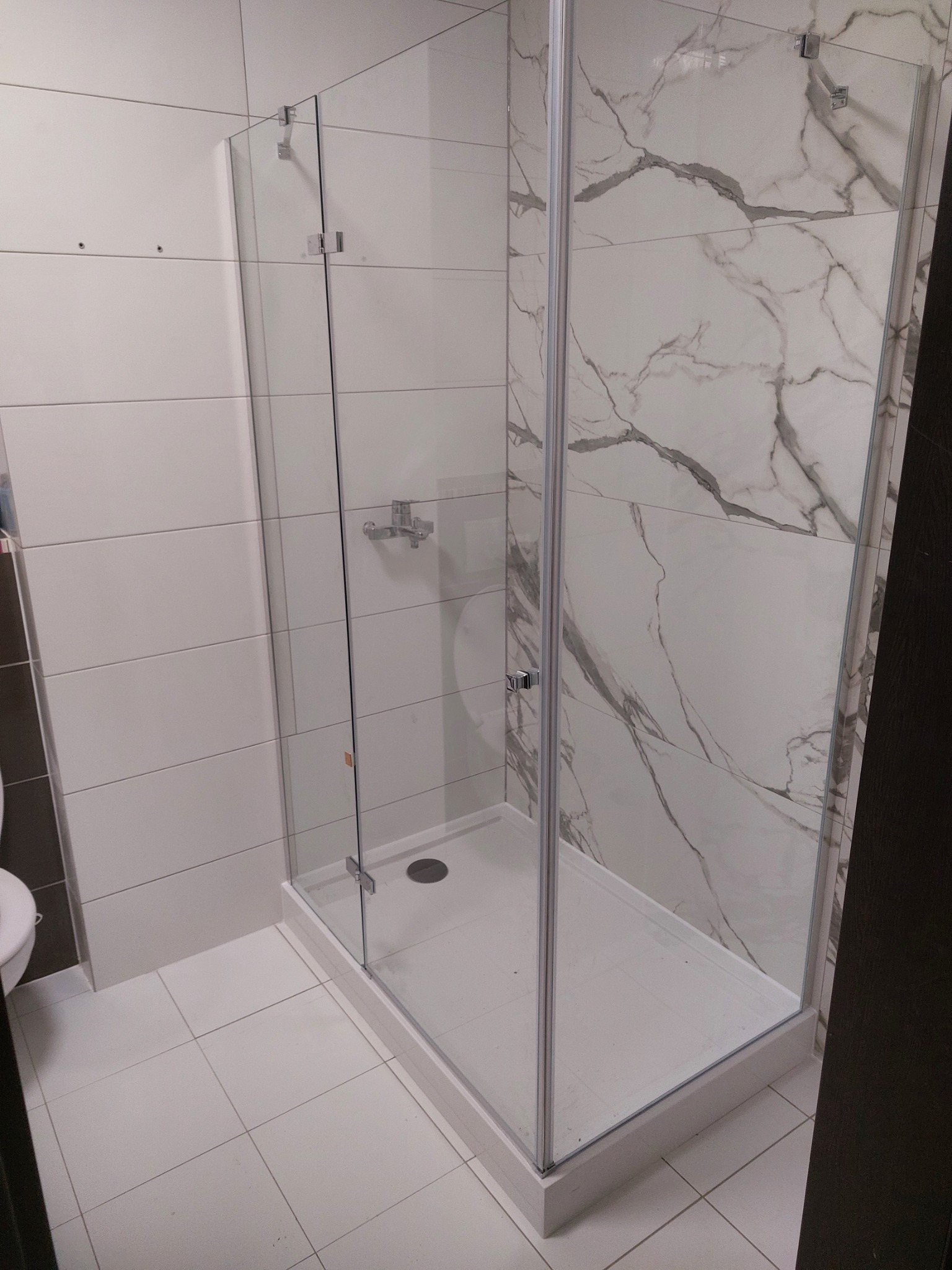 Nowoczesna kabina prysznicowa z marmurową ścianą w Ząbkach. Szklane drzwi z chromowanymi zawiasami, białe płytki podłogowe i minimalistyczna armatura.