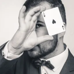 Czarno-białe zbliżenie mężczyzny w garniturze i muszce, zasłaniającego oko asem pik, widoczne mankiety koszuli z ozdobnymi spinkami.